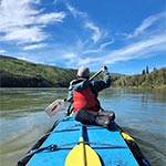 Le dos d'un jeune à l'avant d'un canot à deux places, pagayant le long du fleuve Yukon sous un beau ciel bleu
