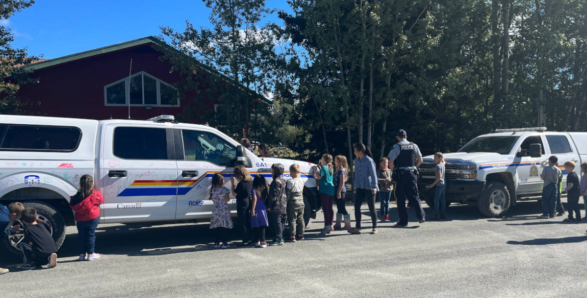 A group of school children colourfully drawing on two police vehicles using dry erase markers as part of the Colouring a Police Vehicle event as community RCMP officers and teachers supervise them.