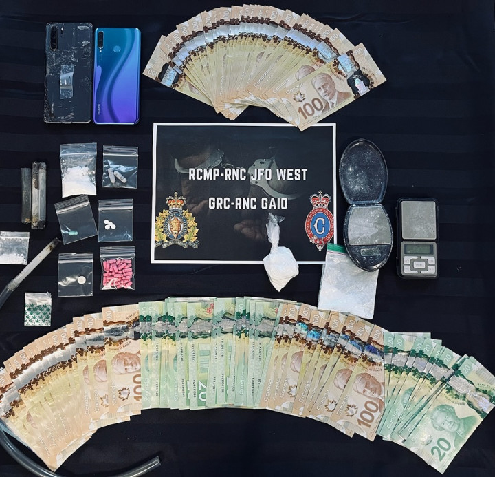 A number of items are displayed on a table including quantities of cash, drugs, prescription pills, cell phones, digital scales and various other items consistent with drug trafficking. A sign reads 